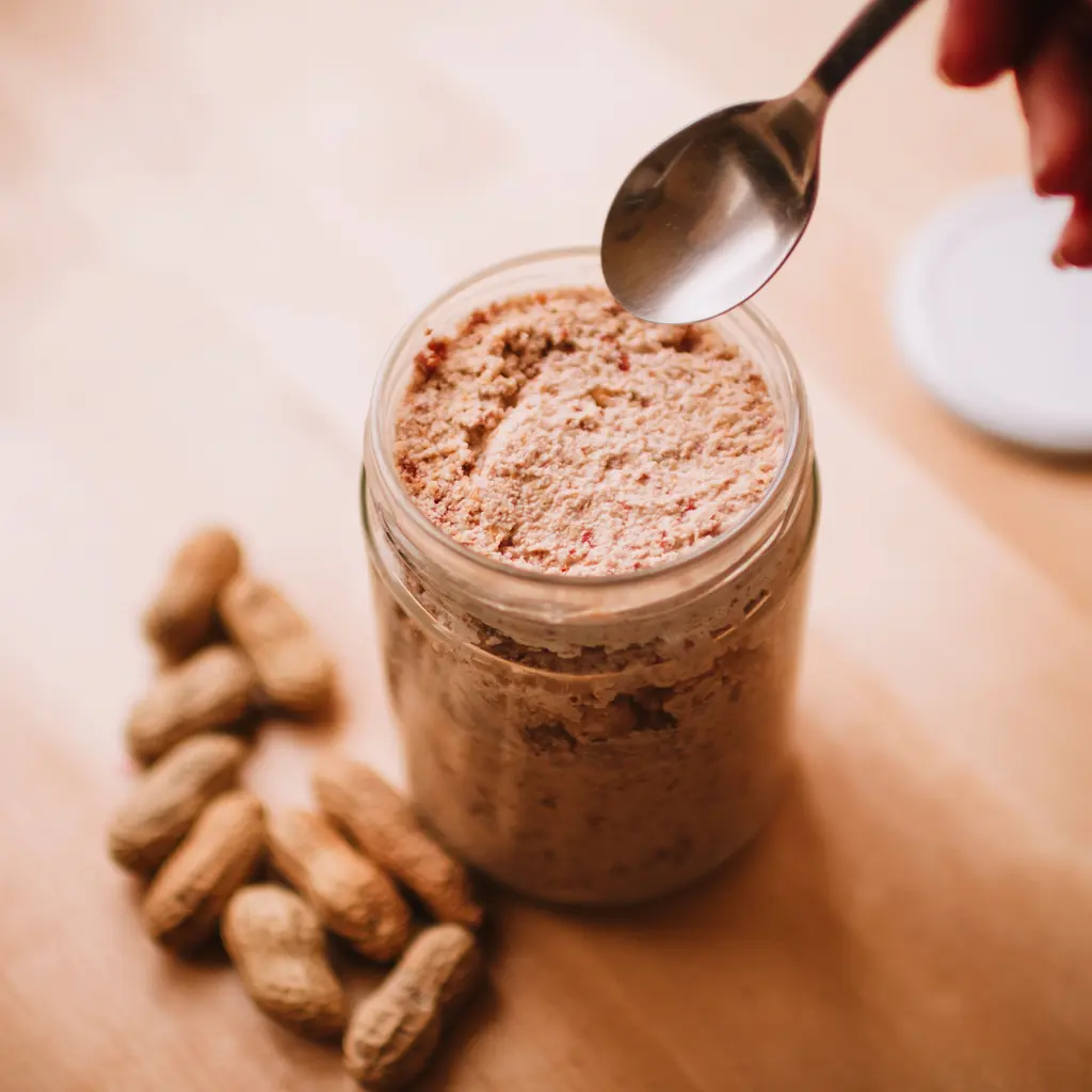 A jar of peanut butter beside whole peanuts and a spoon.