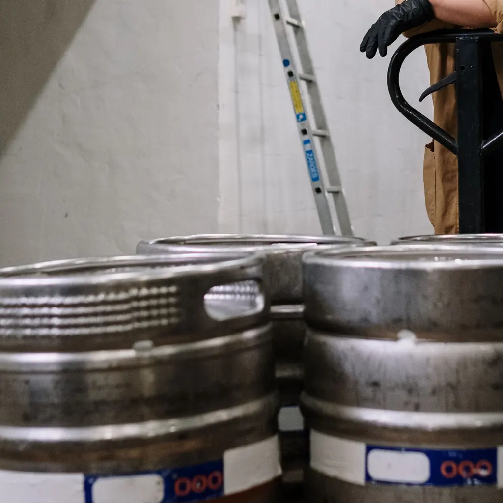 Metal beer kegs and a pallet jack in a brewery storage aisle.