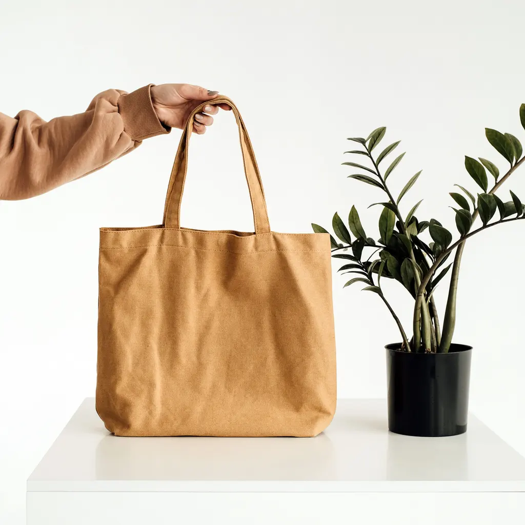 A plain tan canvas tote bag held by its handles beside a potted plant.