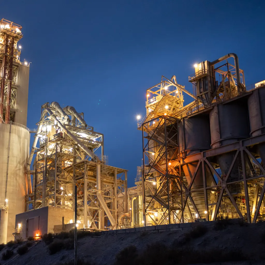 Illuminated towers, silos and processing structures at a cement plant at dusk.