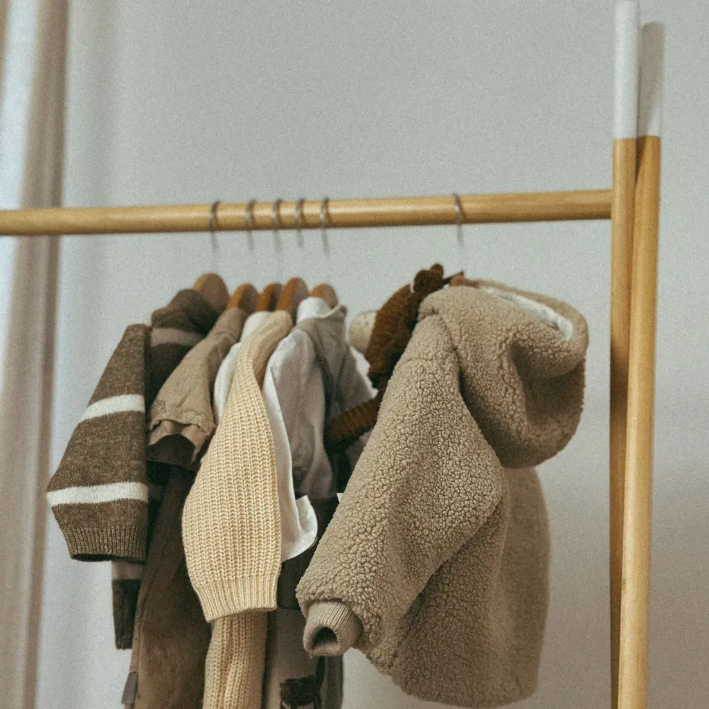 Small children's jackets and knitted sweaters hanging on a wooden clothing rail.