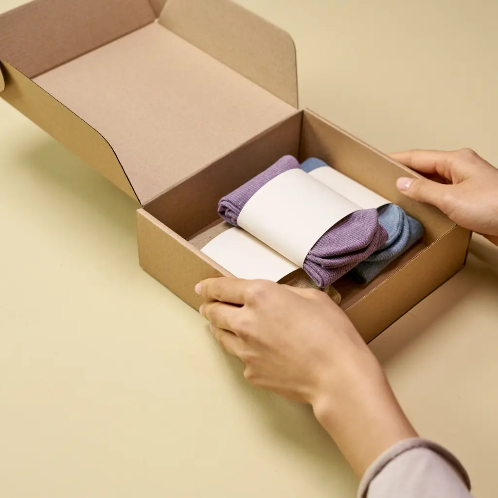 Hands placing folded socks with plain paper bands into a cardboard package.