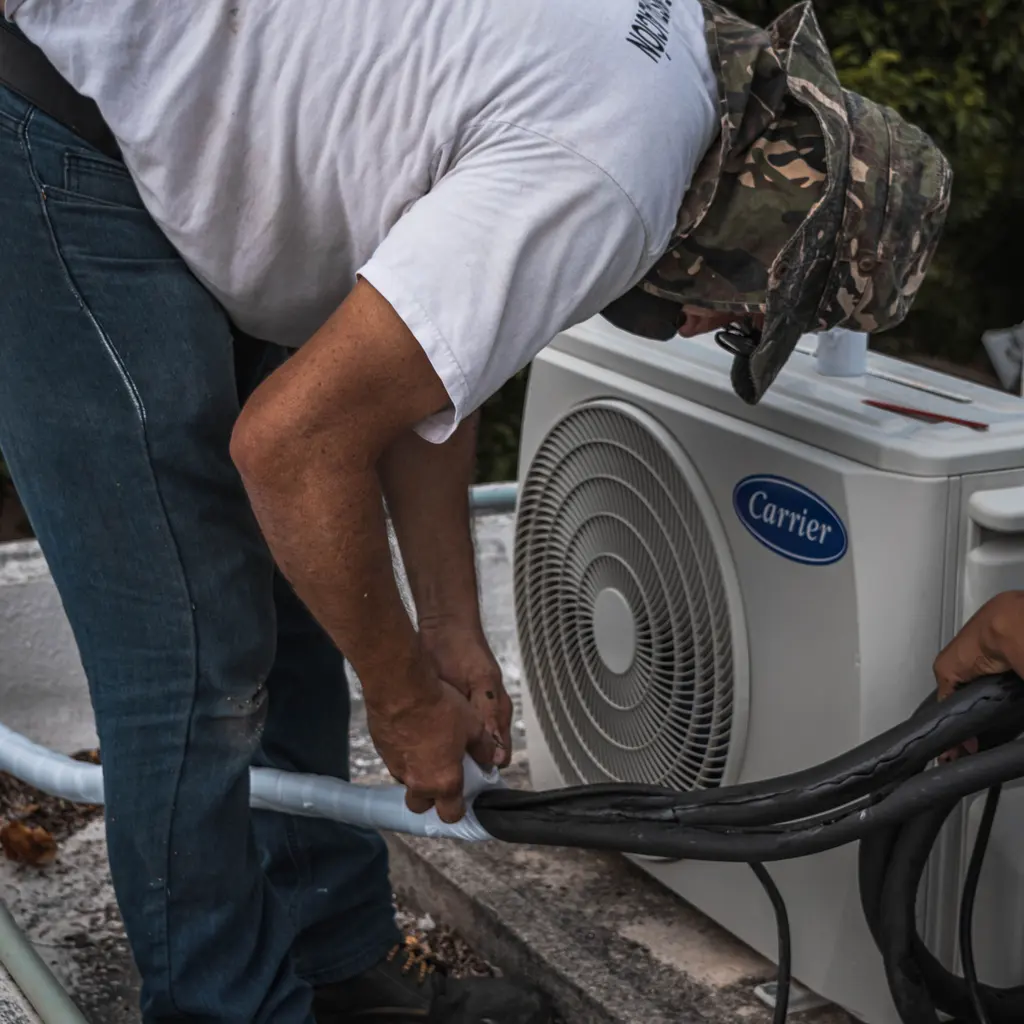 A technician connecting hoses beside an outdoor air-conditioning condenser unit.