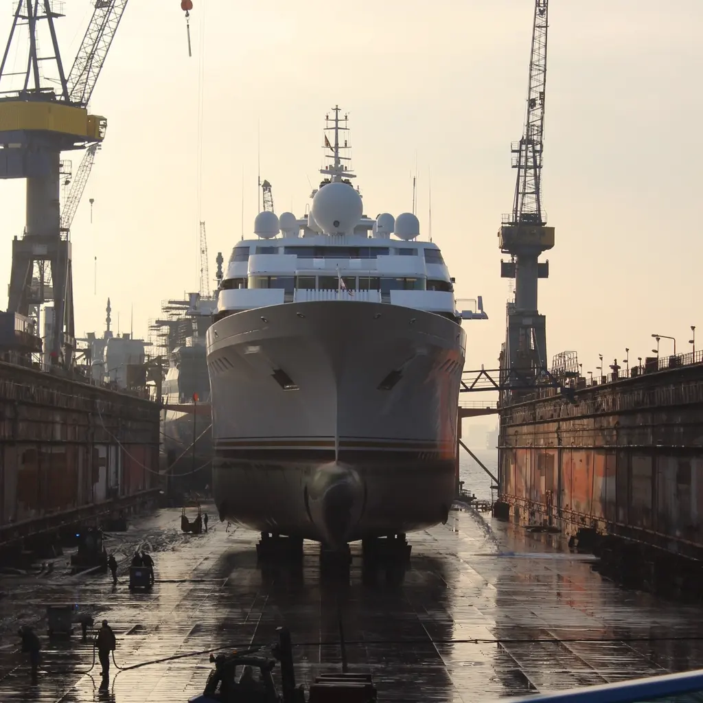 A large yacht supported in a drained shipyard dry dock between cranes and dock walls.