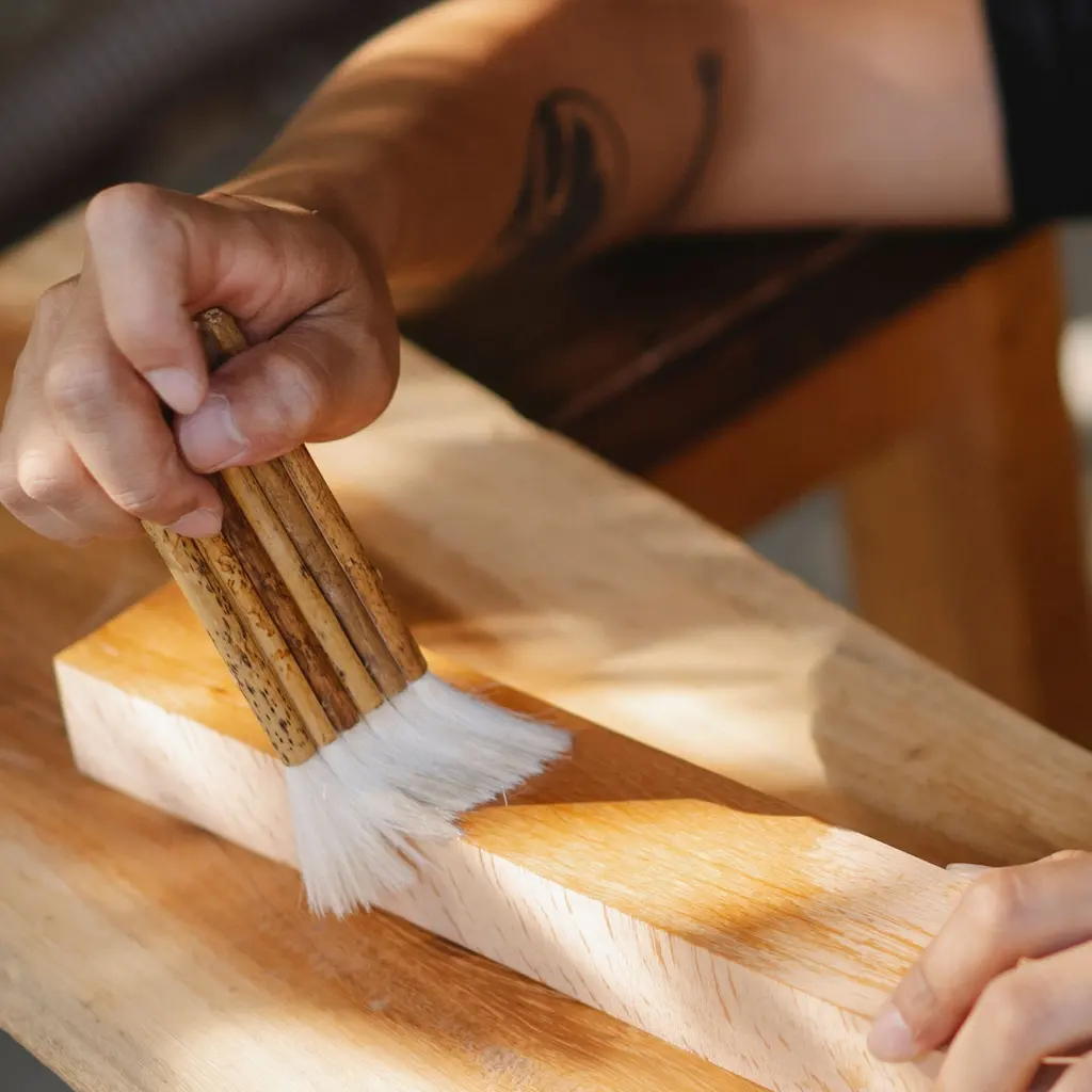 A hand using a wide brush to apply clear varnish to a wooden board.