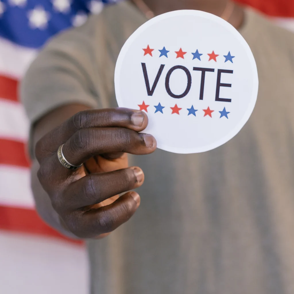 A hand holding a Vote sticker with an American flag behind it.