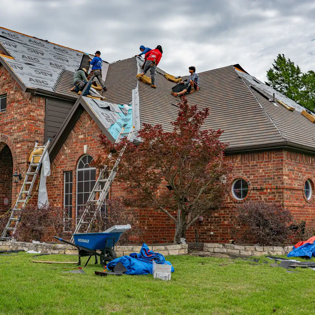 Roofing crew replacing shingles and exposed underlayment on a brick house.