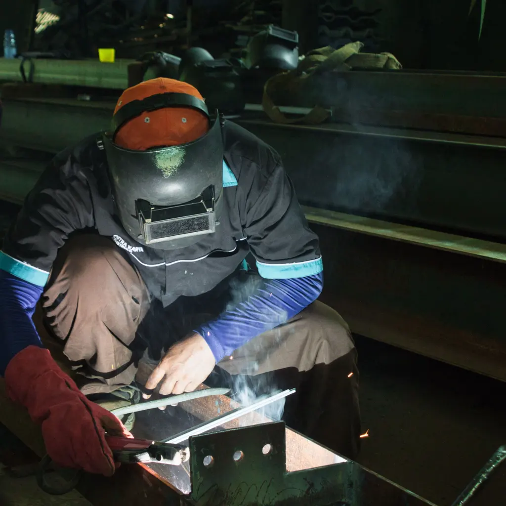 Welder in a protective hood joining a structural steel member inside a fabrication workshop.