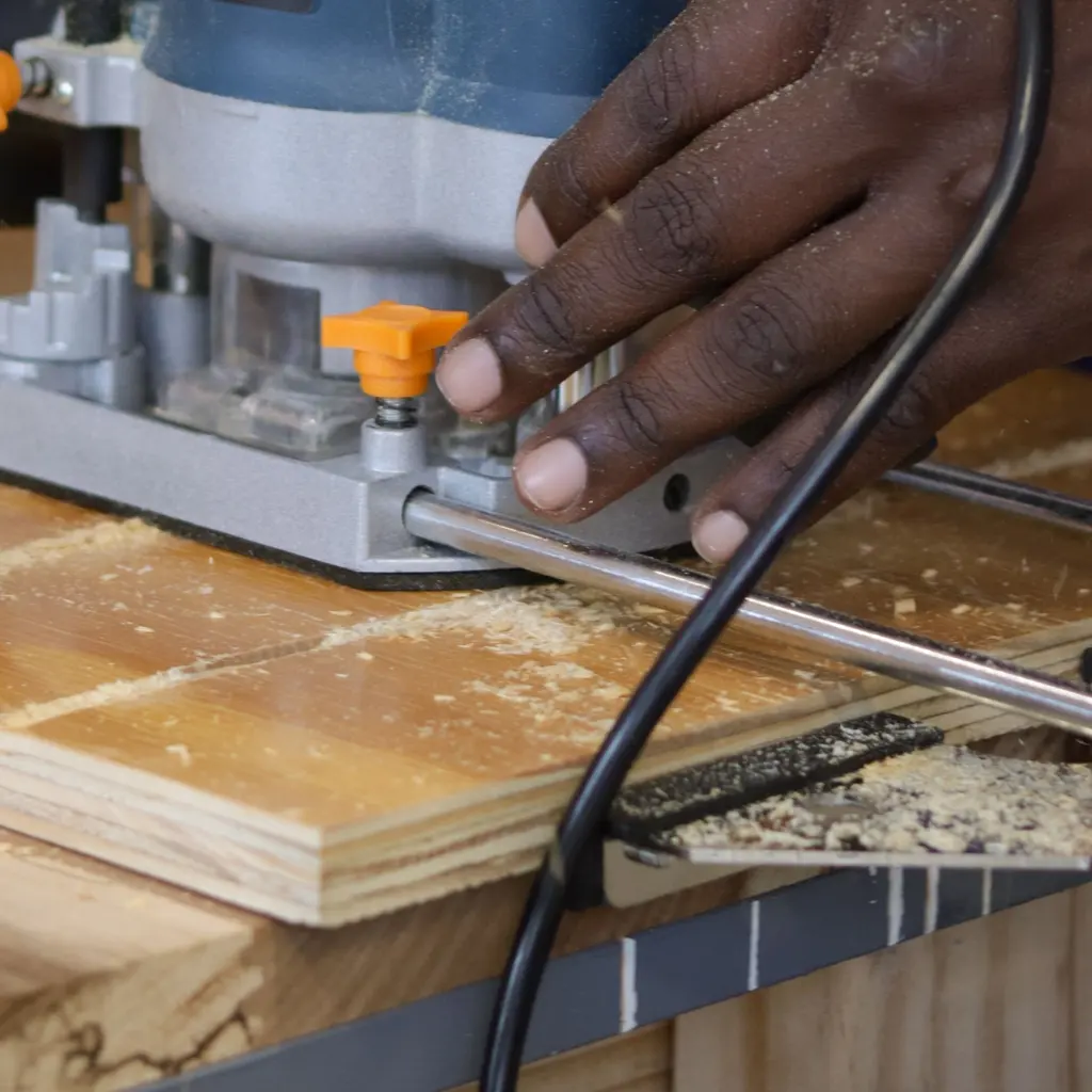 A worker guides a router over a plywood panel on a workbench.