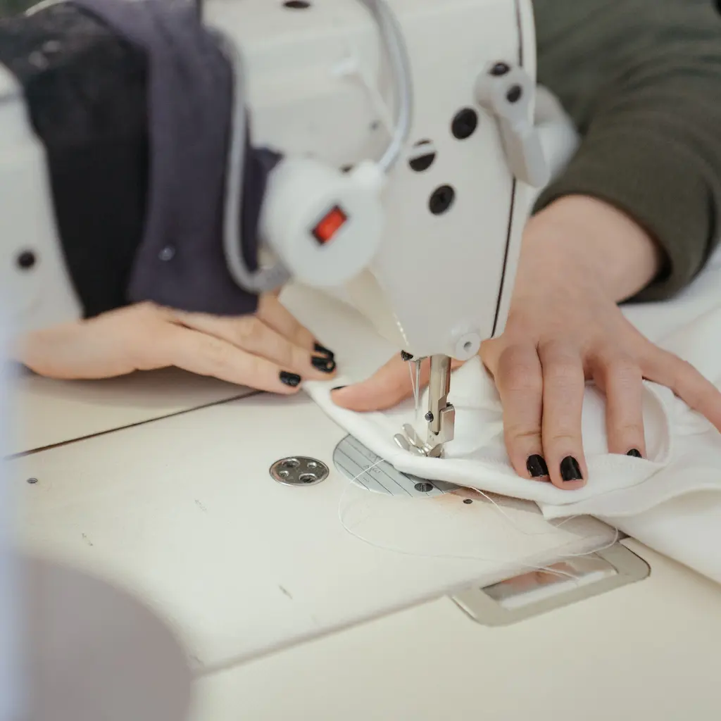 Hands guide white fabric beneath the needle of an industrial sewing machine.