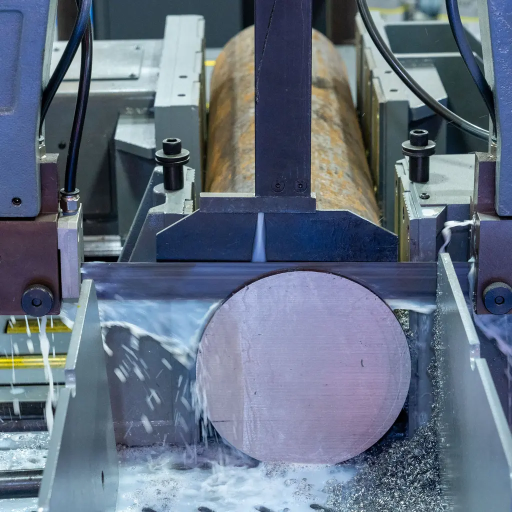 An industrial bandsaw cuts a thick metal bar as coolant runs over the blade and stock.