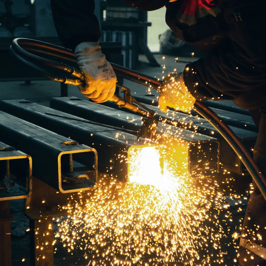 Gloved hands gas-cutting rectangular metal stock in a workshop.