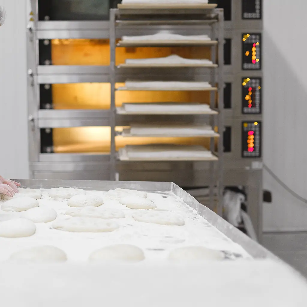 Dough portions on a bakery worktable beside a tray rack and illuminated deck ovens.