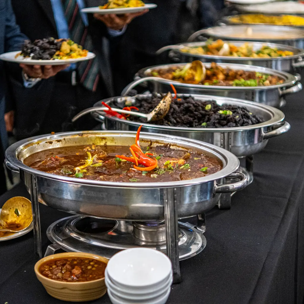 Guests hold plates beside a catered buffet of food in portable stainless-steel chafing dishes.