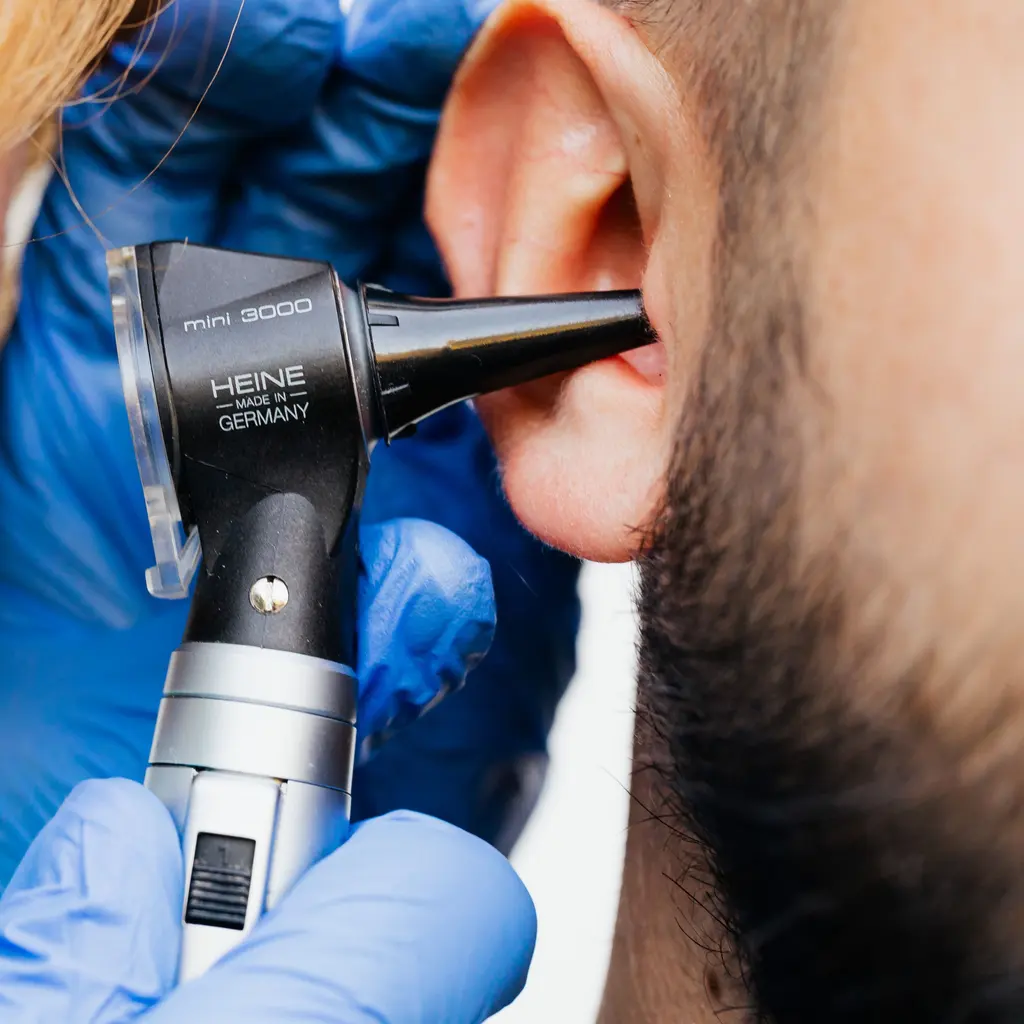 A gloved hand holds an otoscope at an ear during an examination.