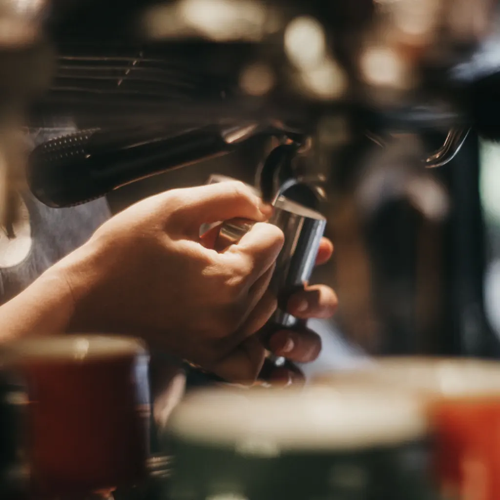 A barista holds a small milk pitcher beneath a commercial espresso machine's steam wand.