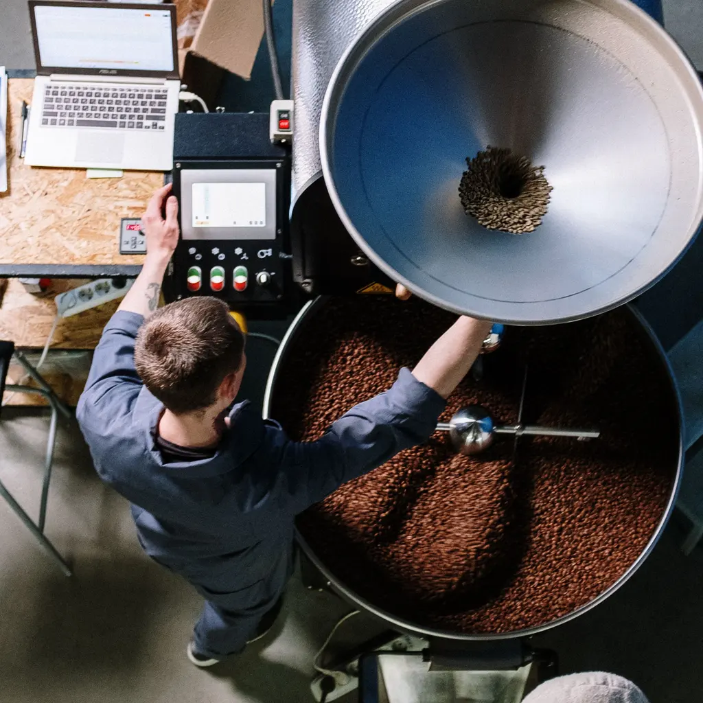 Viewed from above, a worker operates a coffee roaster beside its bean-filled cooling tray.