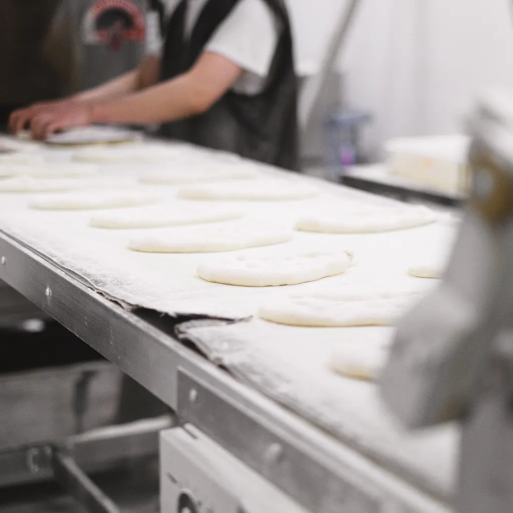 Workers shape rows of dough on a stainless-steel bakery production conveyor.
