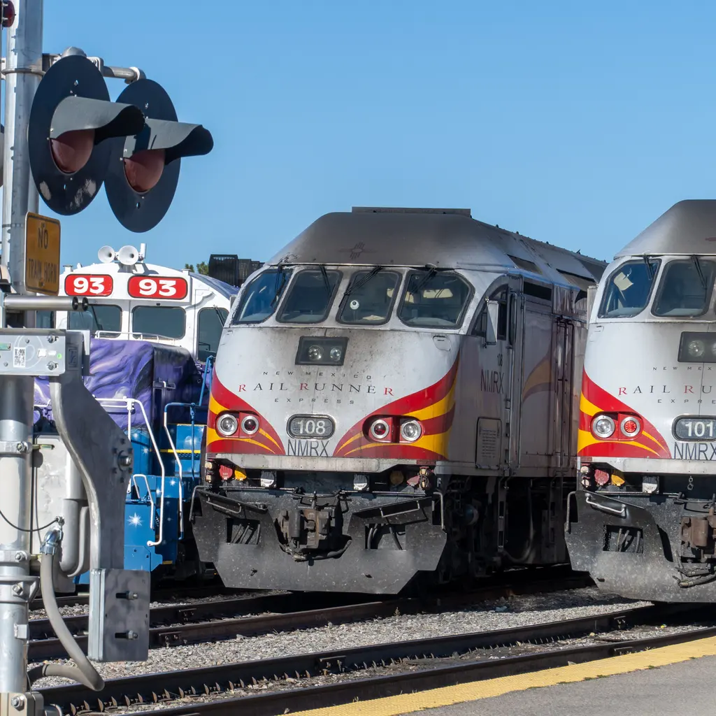 A Rail Runner locomotive stands on yard tracks beside another train and crossing signals.