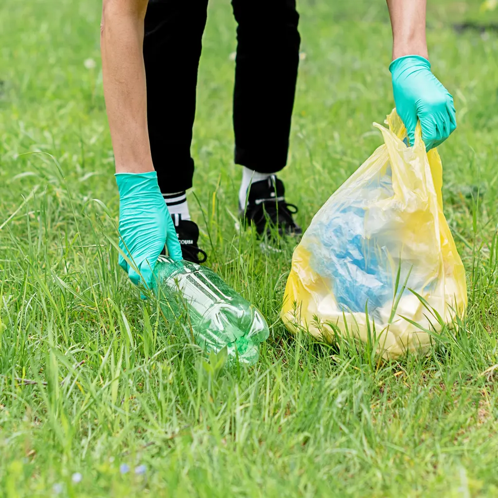 Gloved hands hold a plastic bottle and a litter bag during a park cleanup.