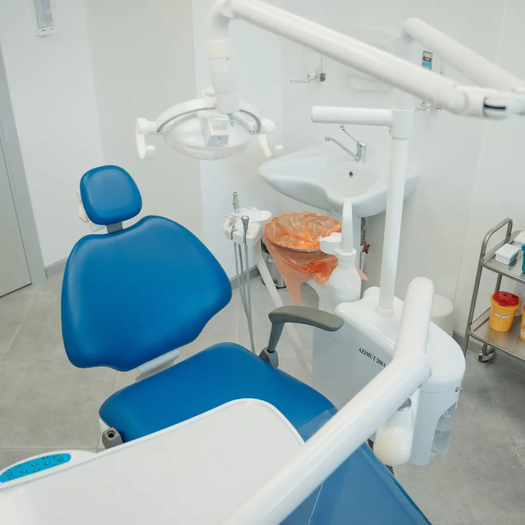 A blue dental chair sits beneath an examination light beside attached handpieces.