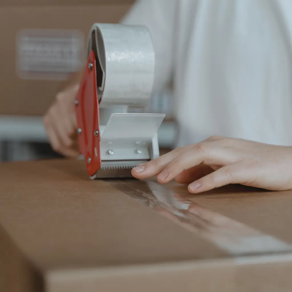 Hands sealing a cardboard parcel with a tape dispenser at a packing station.