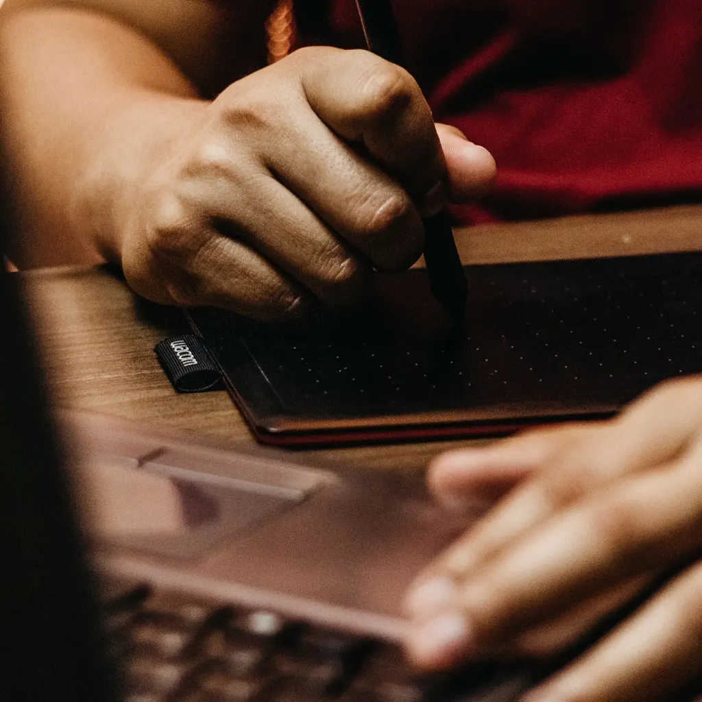 Hands work with a stylus tablet beside an open laptop.
