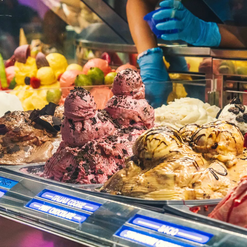 A worker's gloved hands serve from a refrigerated gelato display with labeled flavors.