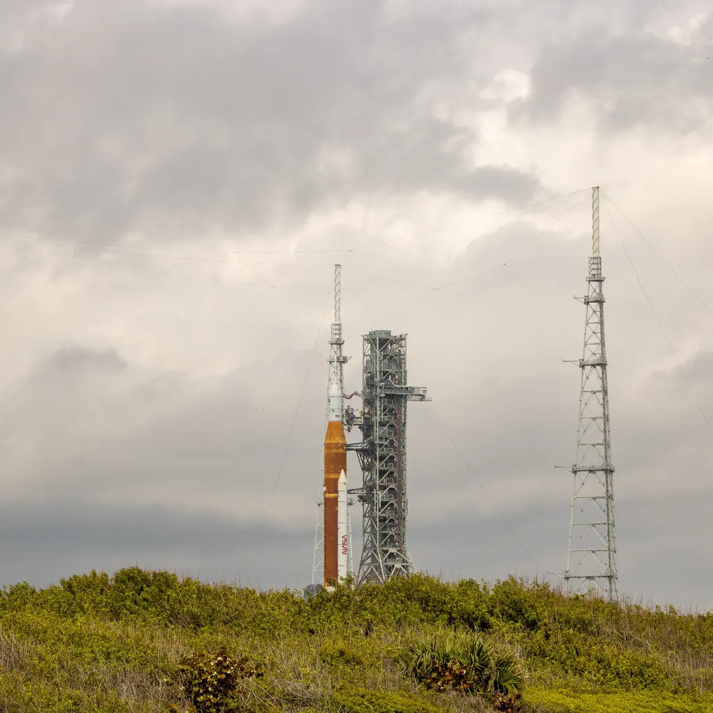 A rocket and its service tower stand between tall lightning-protection towers at a launch pad.