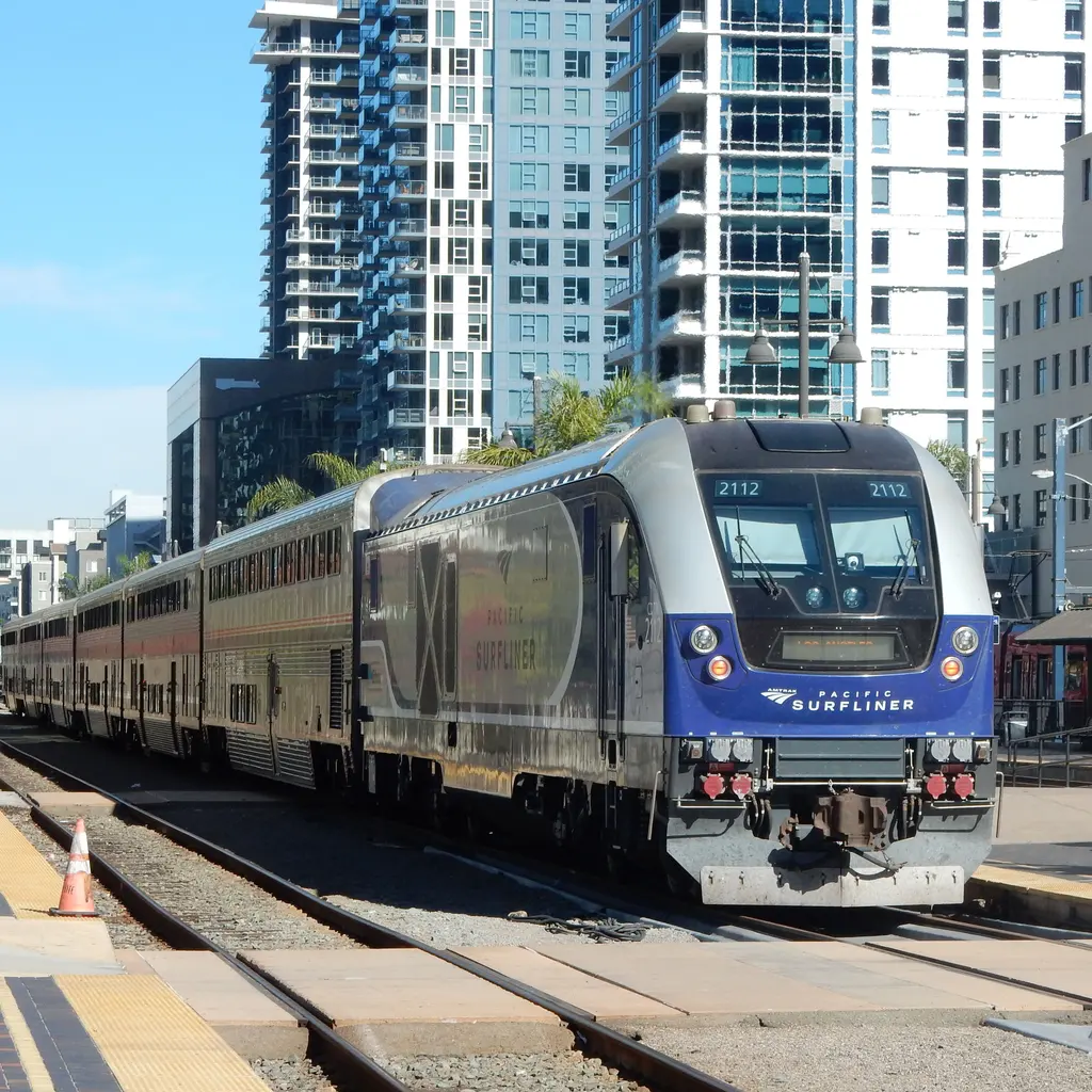 A Pacific Surfliner passenger train sits beside station platforms and a track crossing.