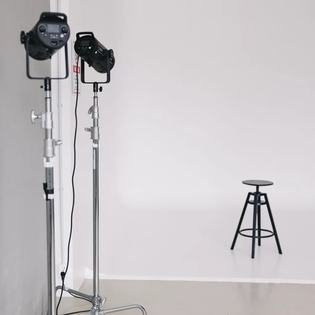 Studio flash lights face a posing stool against a white seamless backdrop.
