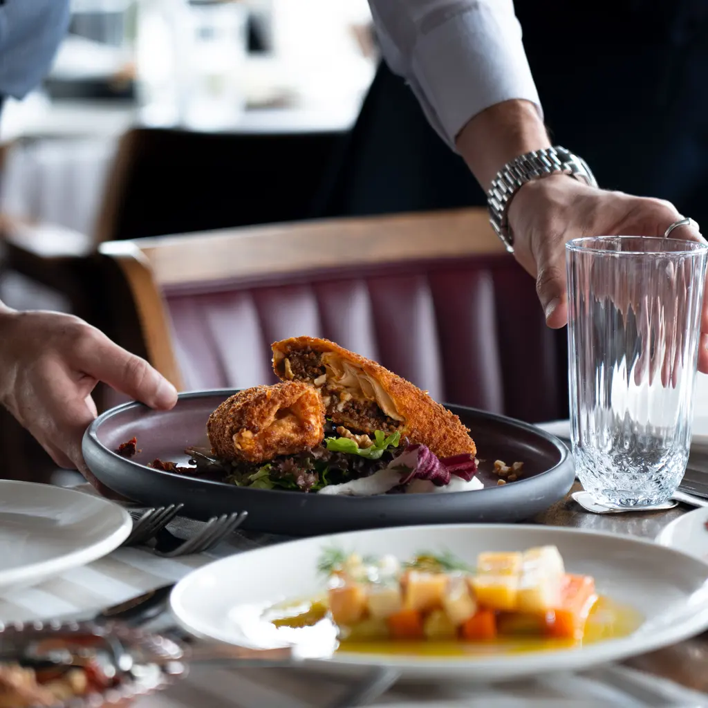 Hands hold a plate of food and a drinking glass at a restaurant table.
