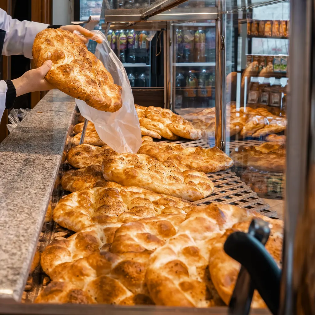 Hands hold a loaf above an open bag beside a bakery bread display.