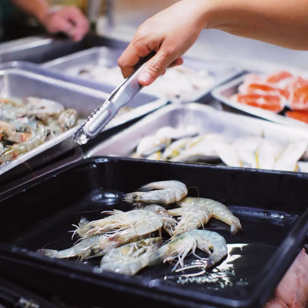 A hand holds tongs above trays of shrimp and fish.