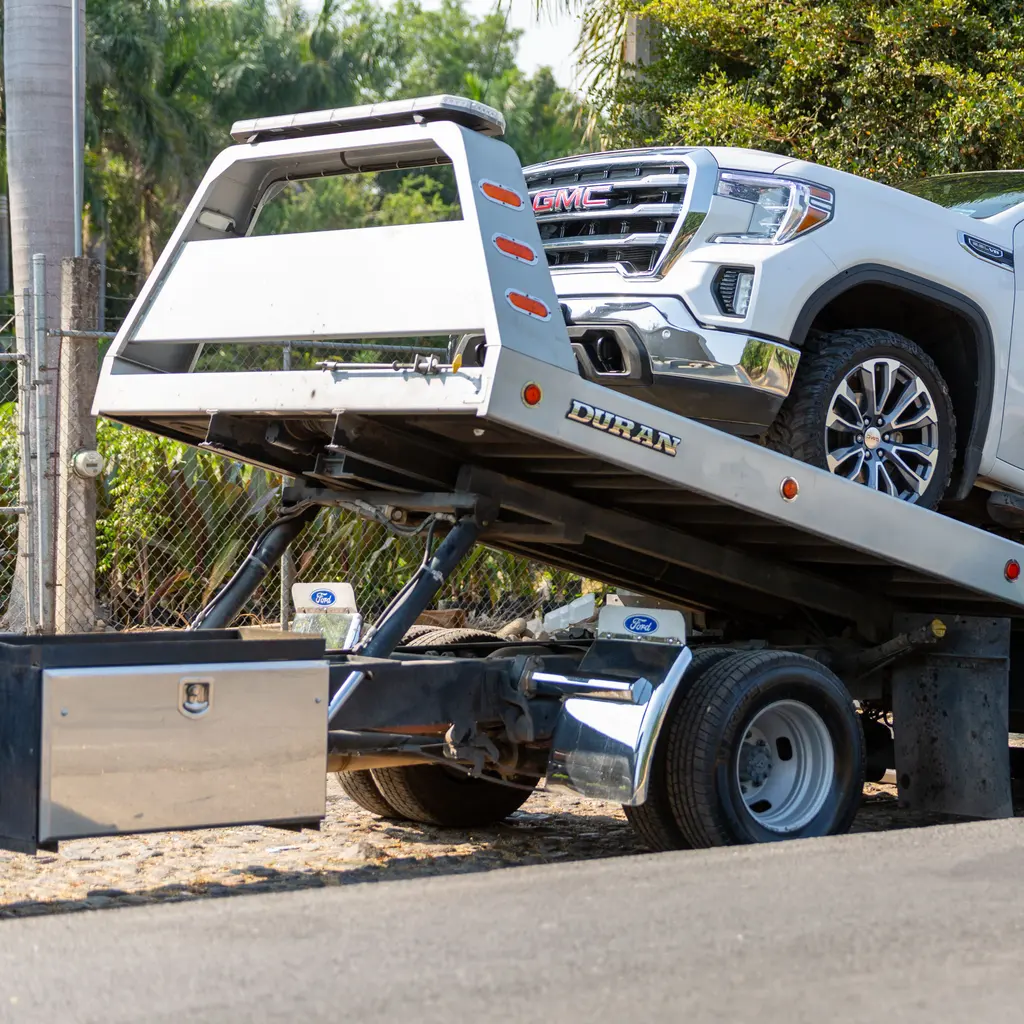 A white pickup rests on a tilted tow-truck bed above its hydraulic lifting mechanism.