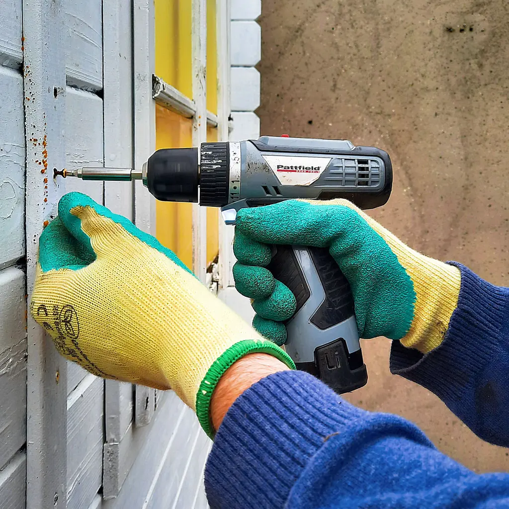 Gloved hands work with a cordless driver at a wooden window-shutter screw.
