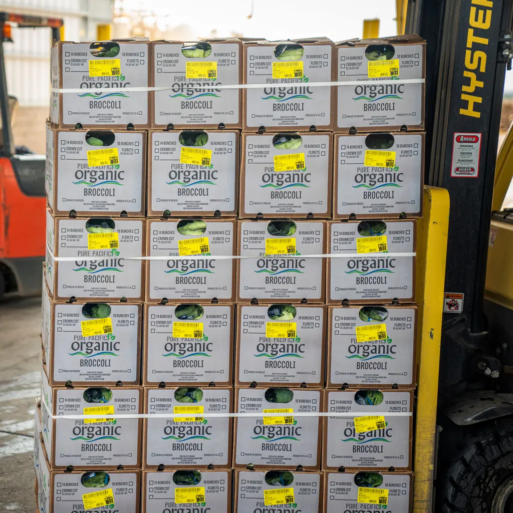 Cartons of broccoli are stacked beside a forklift mast in a produce warehouse.