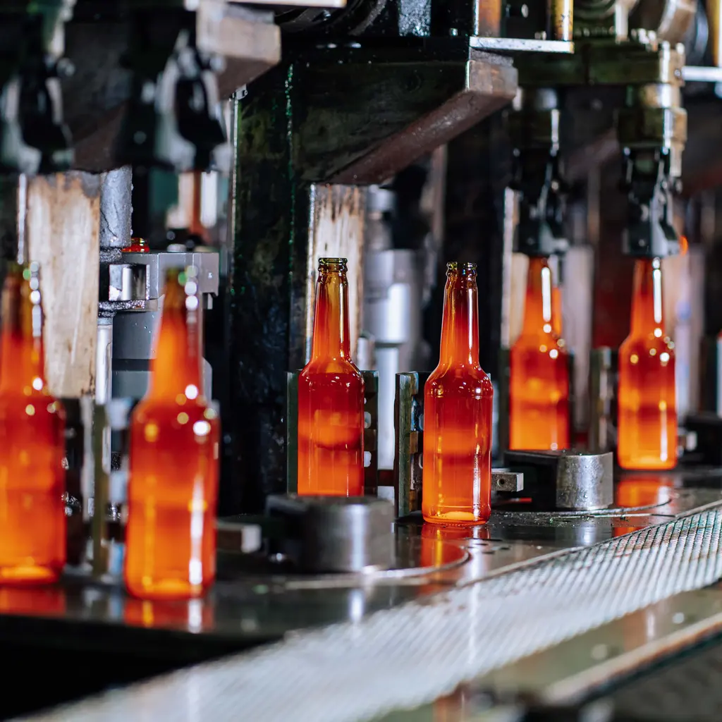 Glowing newly formed glass bottles move between forming machinery on a factory line.