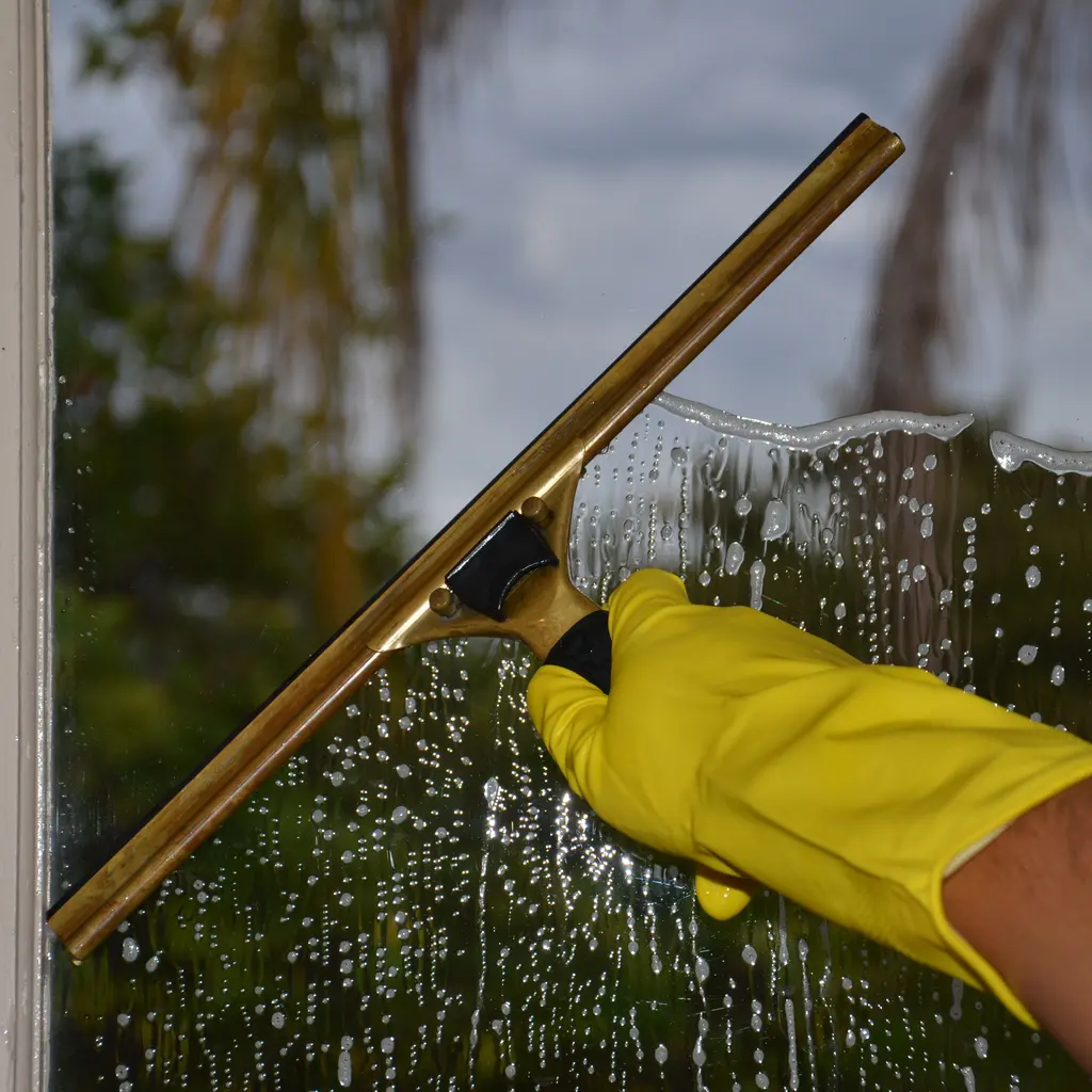 A gloved hand drawing a squeegee across soapy window glass.