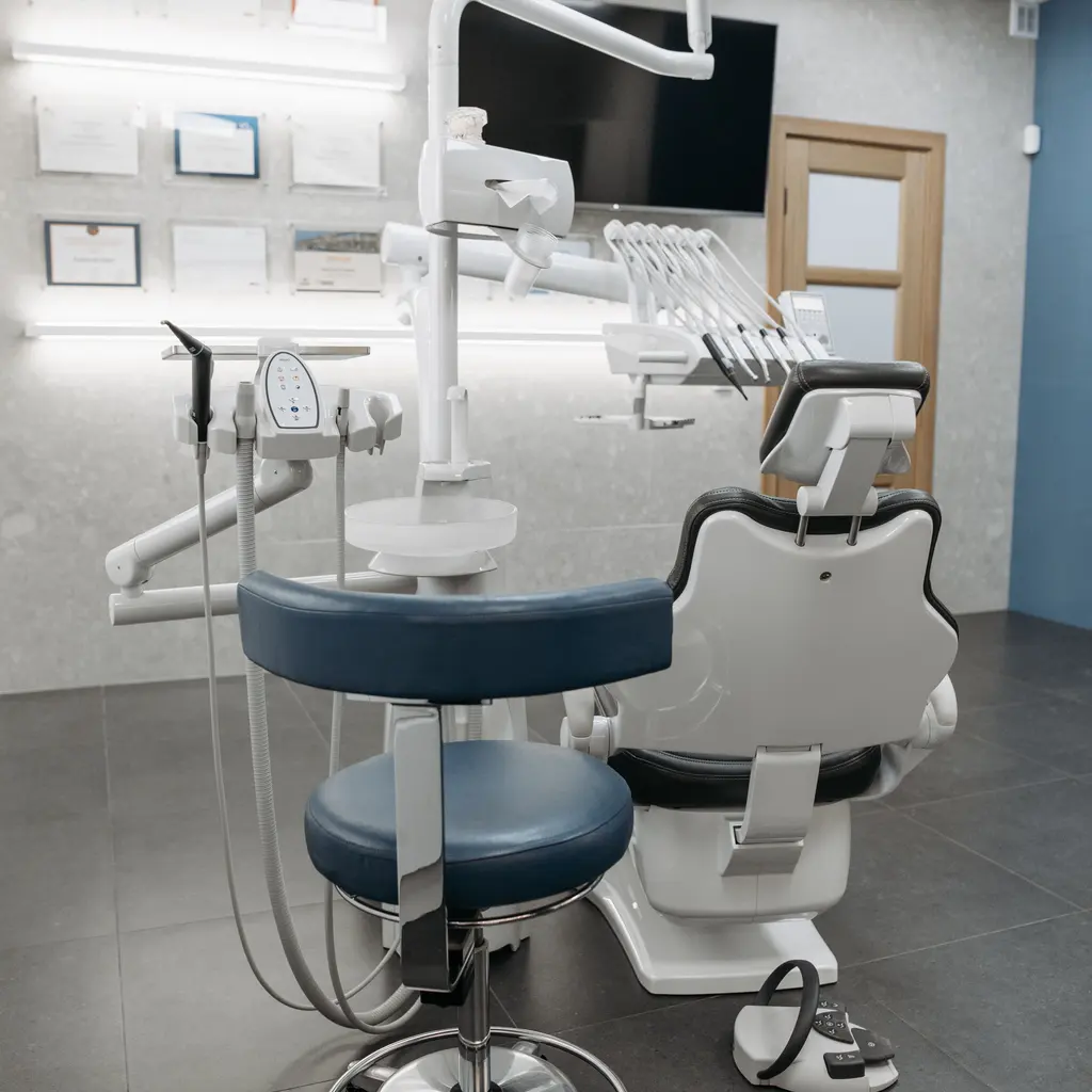An empty dental treatment room with a patient chair, stool and instrument delivery unit.