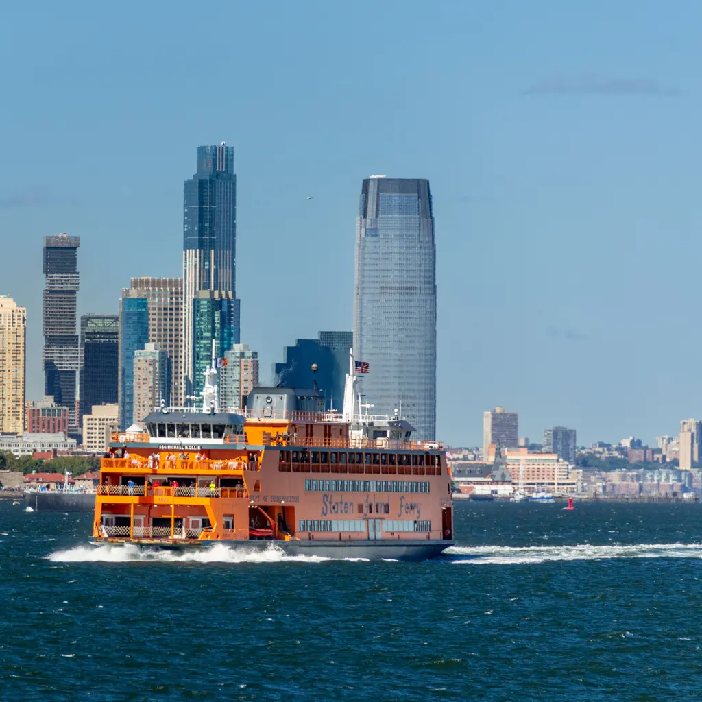 An orange passenger ferry under way with a city skyline behind it.