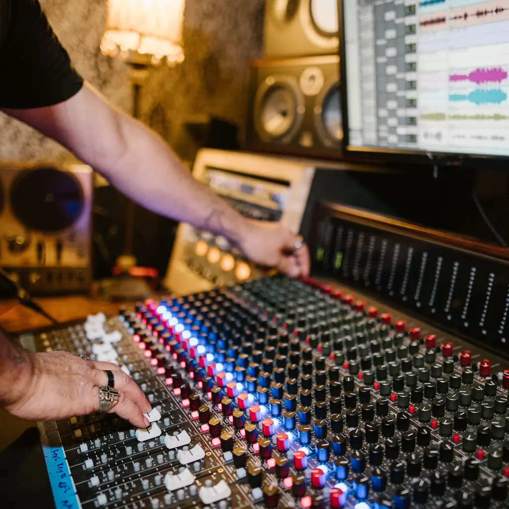 Hands adjusting an audio mixing console beside a multitrack recording display.