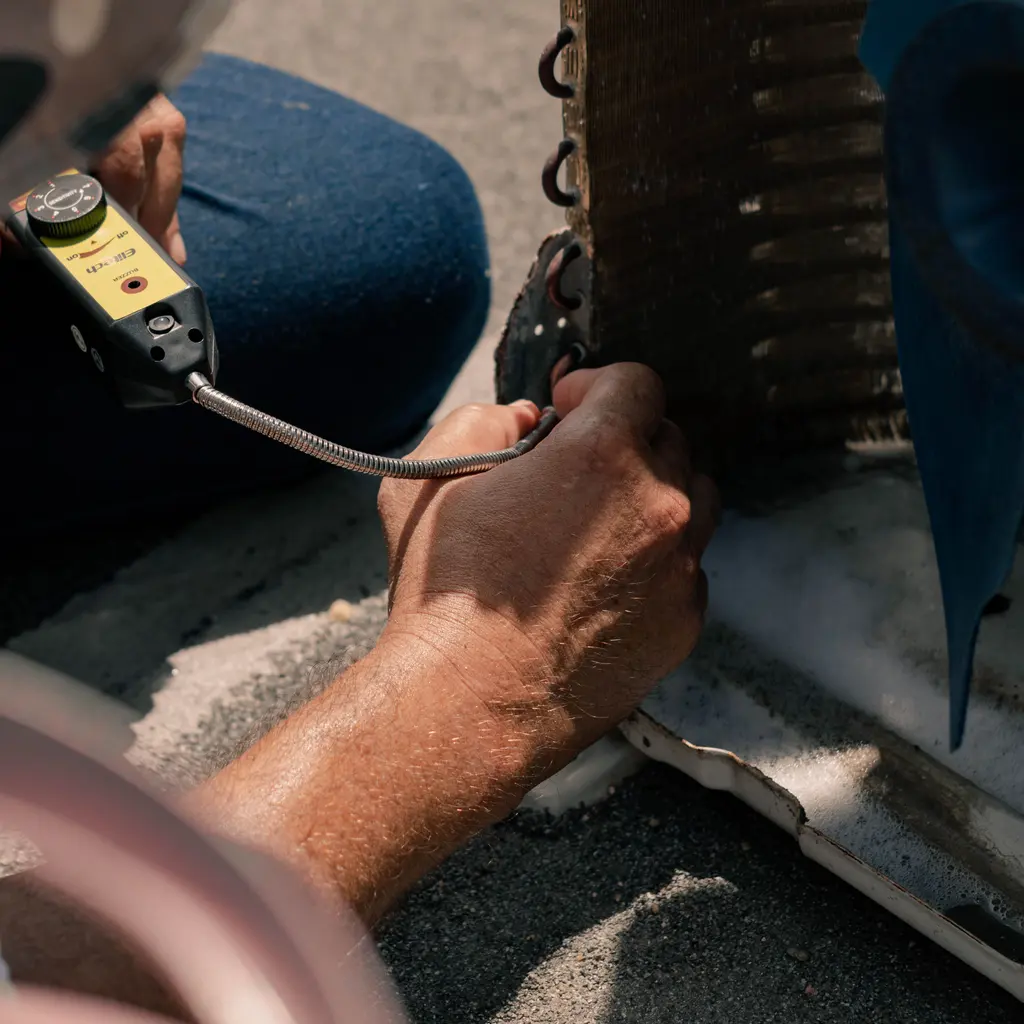 A hand holding a detector probe beside exposed air-conditioning coil tubing.
