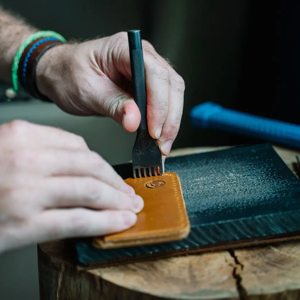 Hands position a stitching chisel along the edge of a leather wallet on a work block.