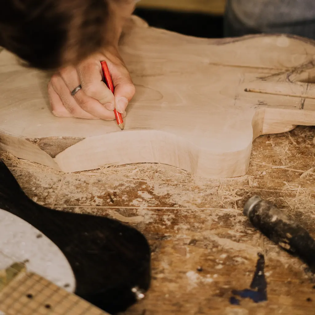 A hand marks an unfinished wooden guitar body with a pencil at a workbench.