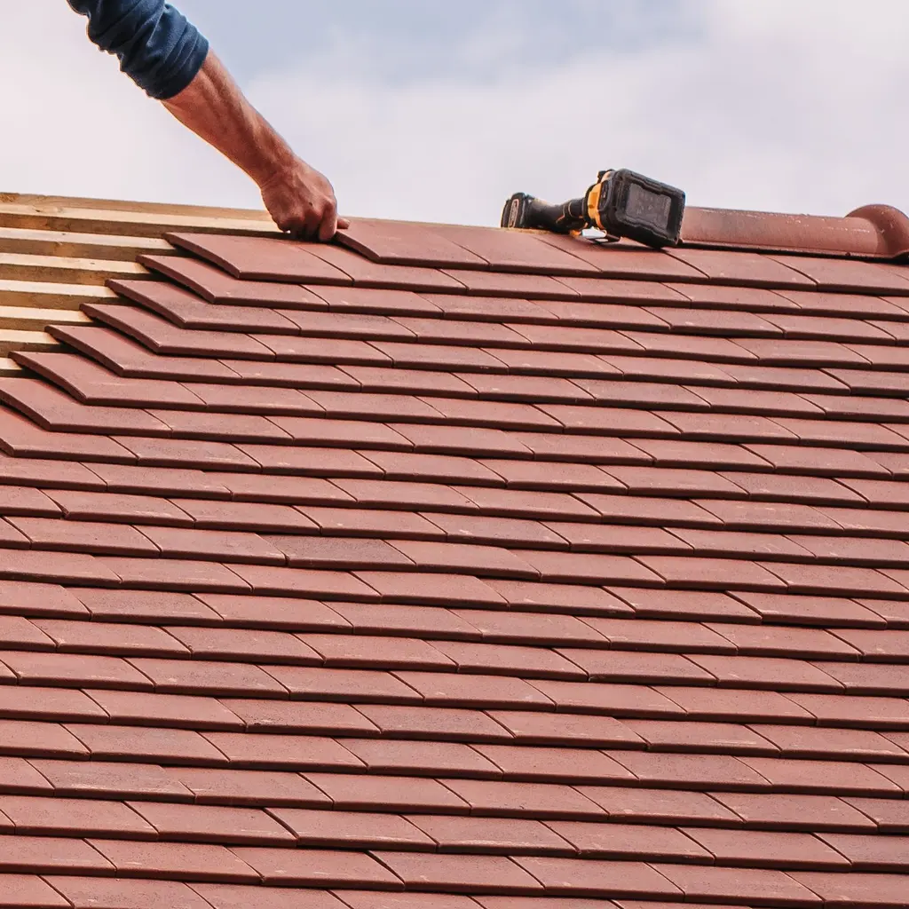 A hand positioning a roof tile beside exposed wooden battens.