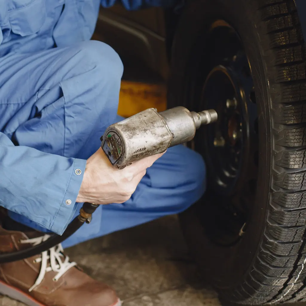 A mechanic works on wheel fasteners with a pneumatic impact wrench inside an auto-service bay.