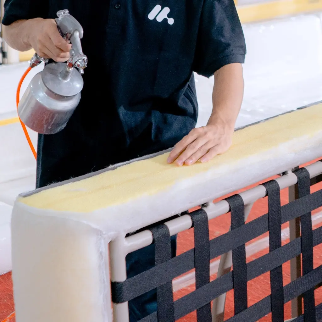 A worker holds a spray gun over foam padding on an unfinished, webbed sofa frame.