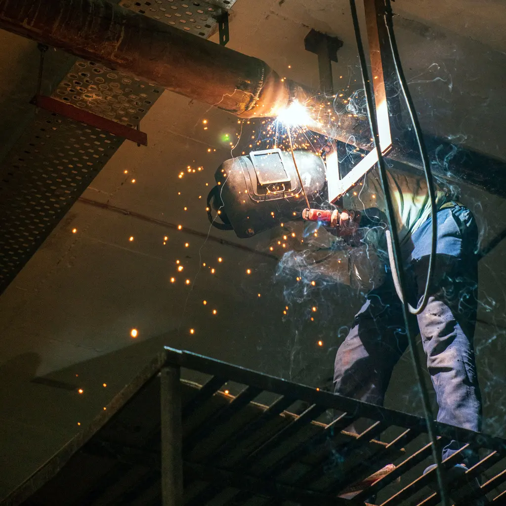A shielded welder works at a metal pipe joint.