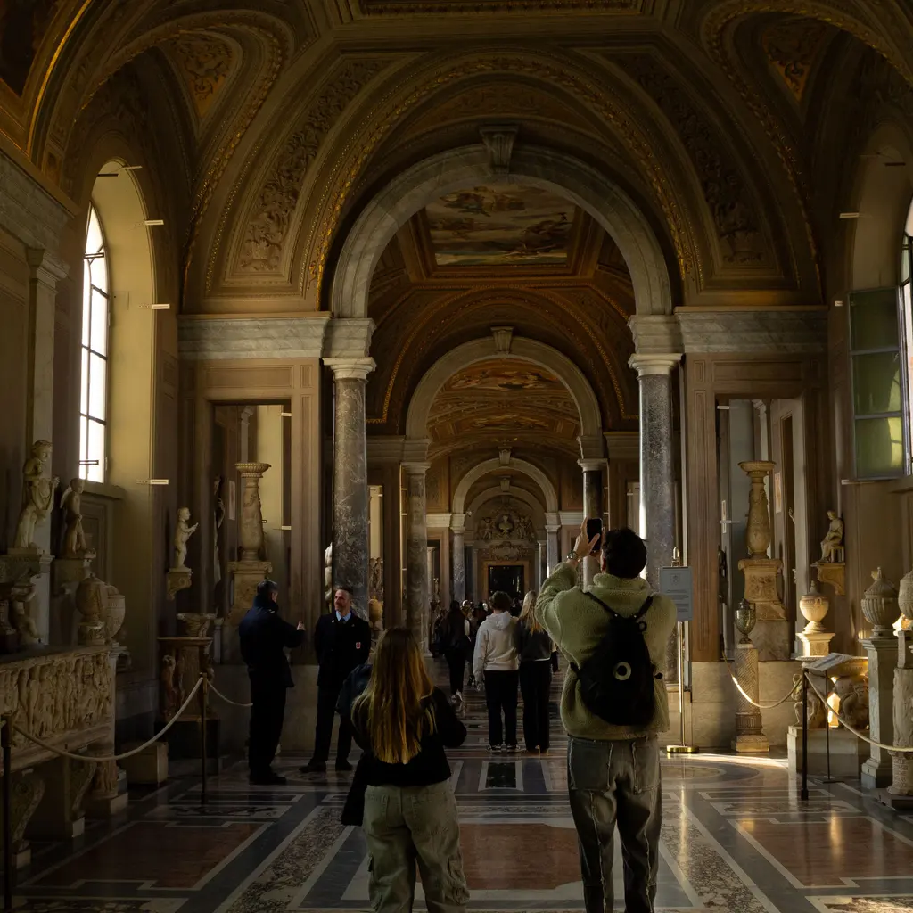 Visitors walking past classical sculptures in a long vaulted museum gallery.